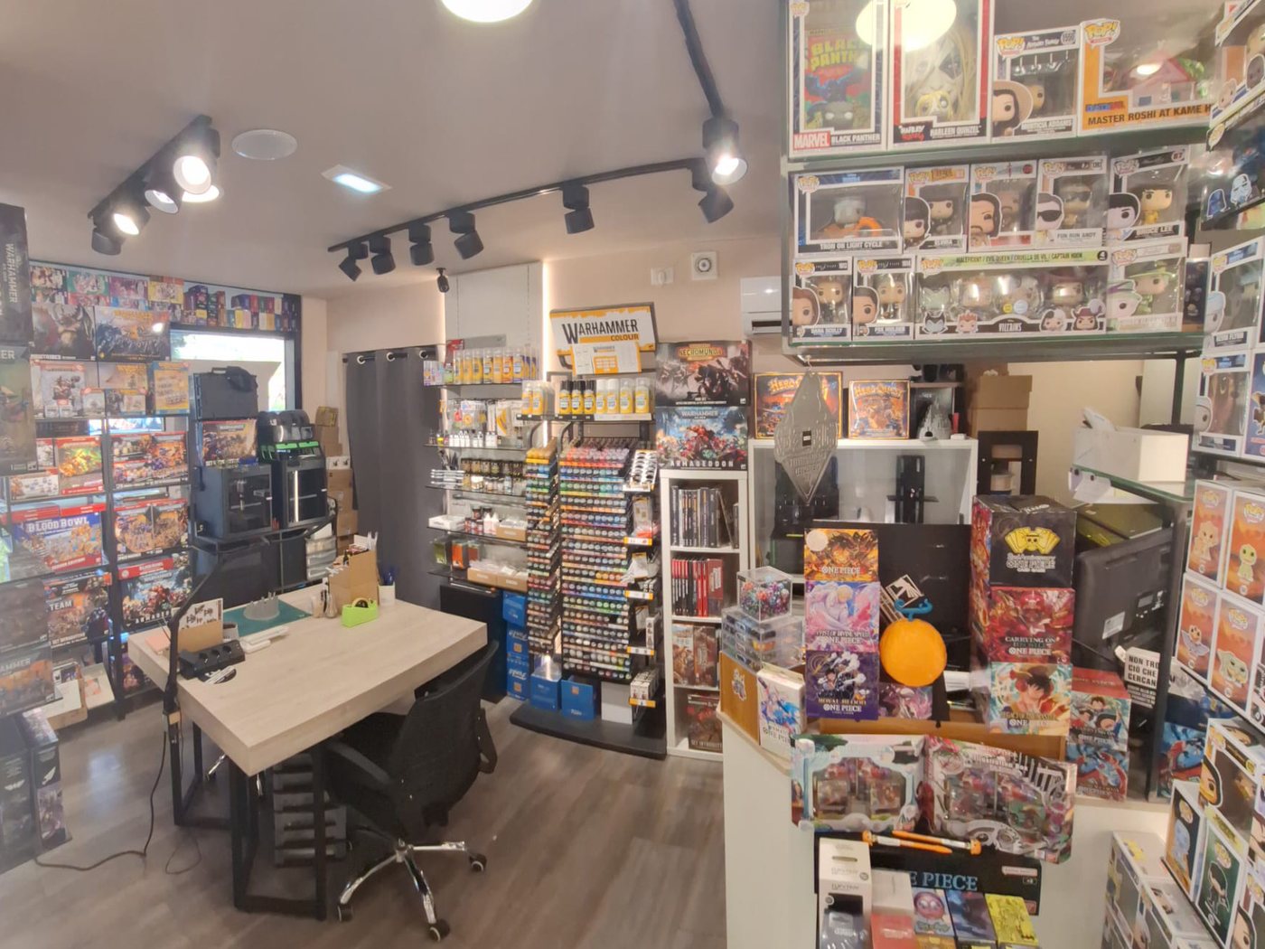 Interno di Forge of GLAM con tavolo in legno chiaro, espositore di colori Warhammer, scatole di giochi e Funko Pop sugli scaffali